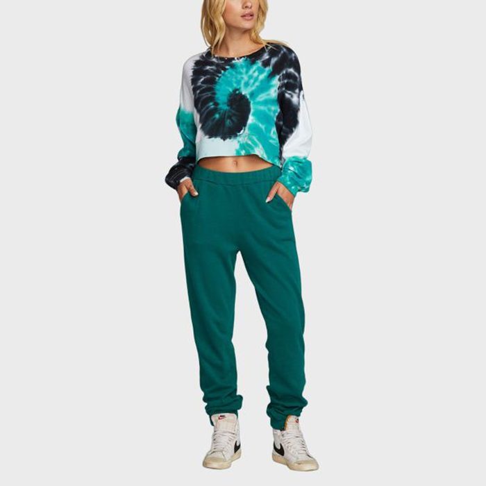 Person wearing tie-dye cropped sweatshirt, teal sweatpants, white sneakers; standing with hands in pockets against a plain light background.