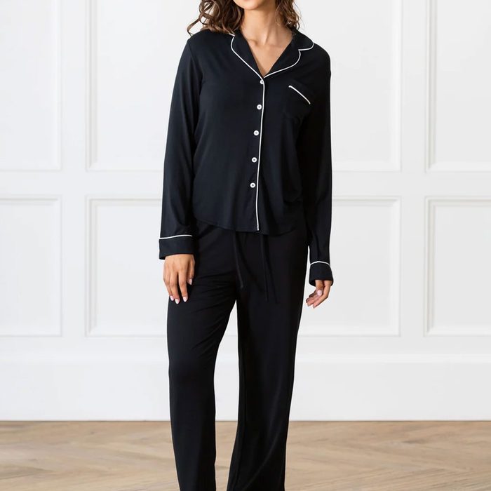 A person wearing a black pajama set stands against a white paneled wall with a wooden floor.