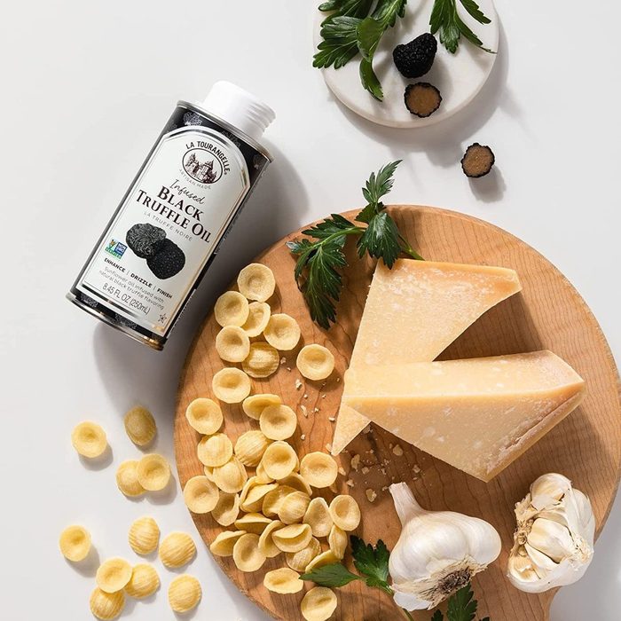 Truffle oil lays next to orecchiette pasta, Parmesan cheese, garlic, and herbs on a wooden board. Black truffle slices decorate a small plate.
