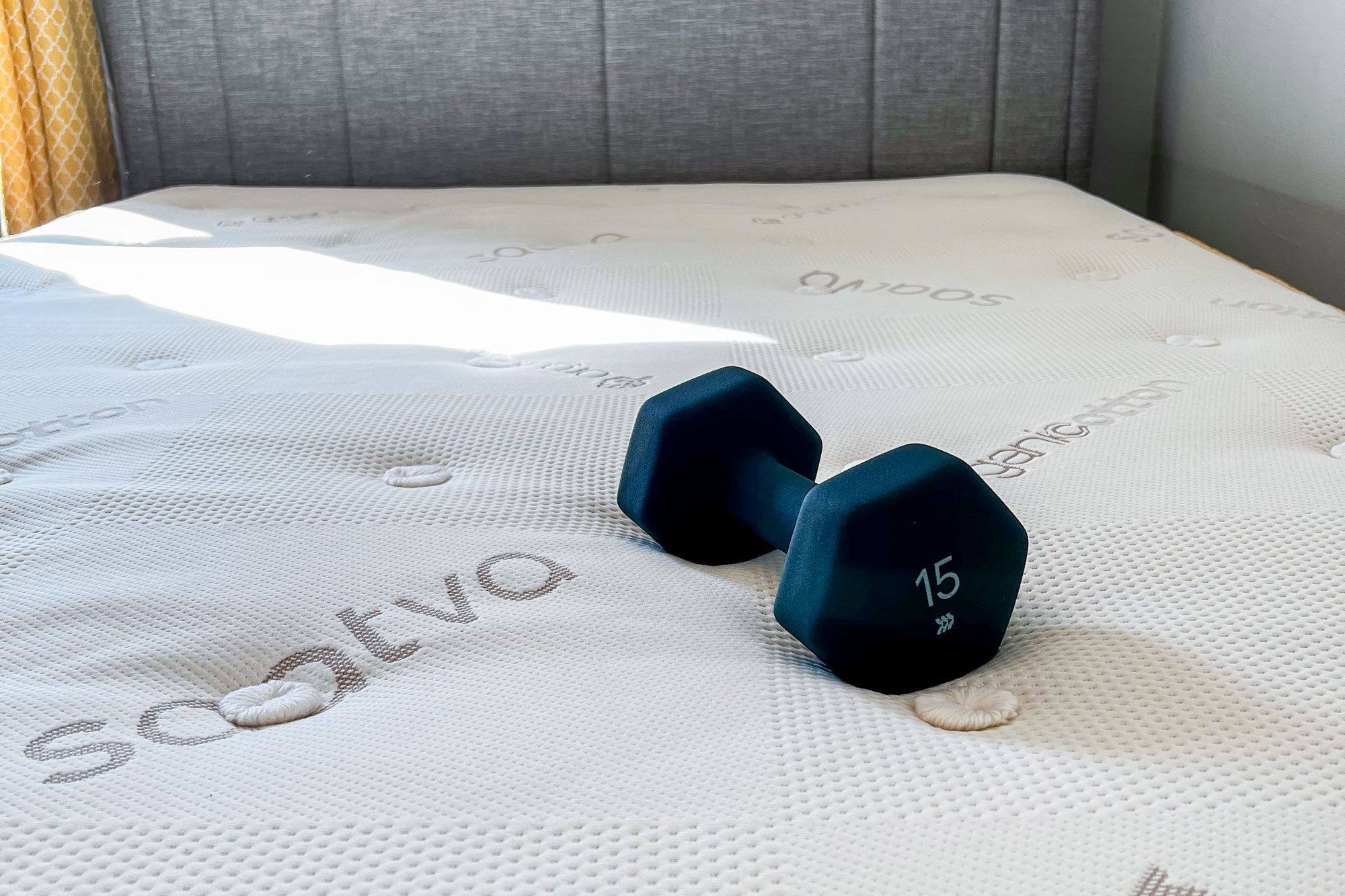 A 15-pound dumbbell rests on a Saatva mattress in a sunlit bedroom with gray headboard and patterned yellow curtains.