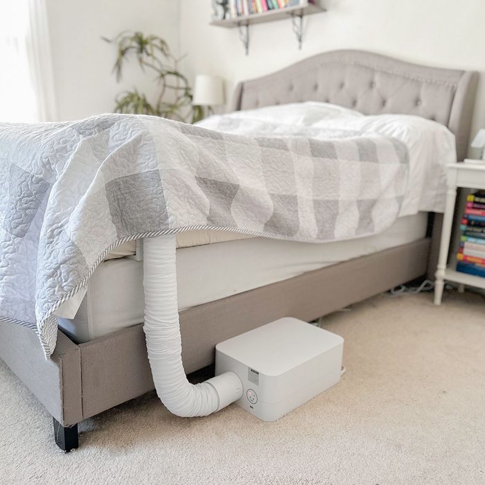 A white device connects via a flexible hose to a bed, situated in a cozy bedroom with a plant and bookshelf nearby.