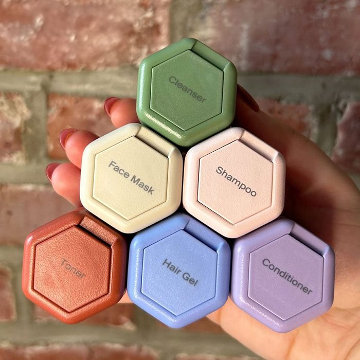 Hand holds hexagonal containers labeled Cleanser, Face Mask, Shampoo, Toner, Hair Gel, Conditioner; set against a brick background.