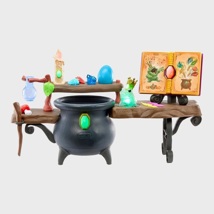 Cauldron sits on wooden table, surrounded by magical items like crystals, a potion bottle, a frog figurine, and an open spellbook.