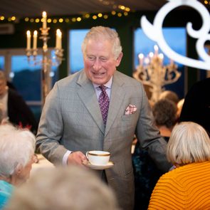 Man in suit holding teacup, smiling while greeting seated elderly people in warmly lit gathering with candelabras and fairy lights.