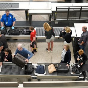 Passengers place luggage on conveyor belt for security screening; a uniformed officer monitors the process in a busy airport terminal.