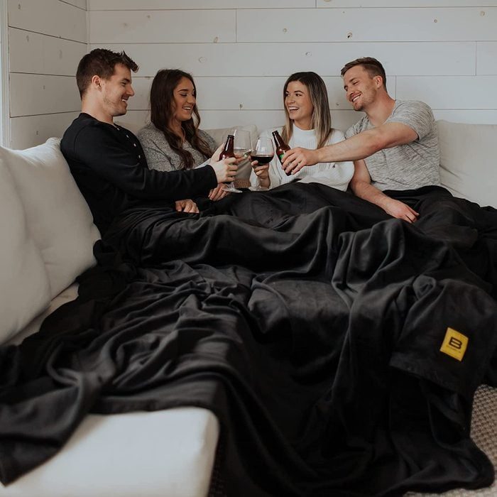 Four people sit on a couch, toasting with drinks, covered by a large black blanket in a cozy room with white wooden walls.