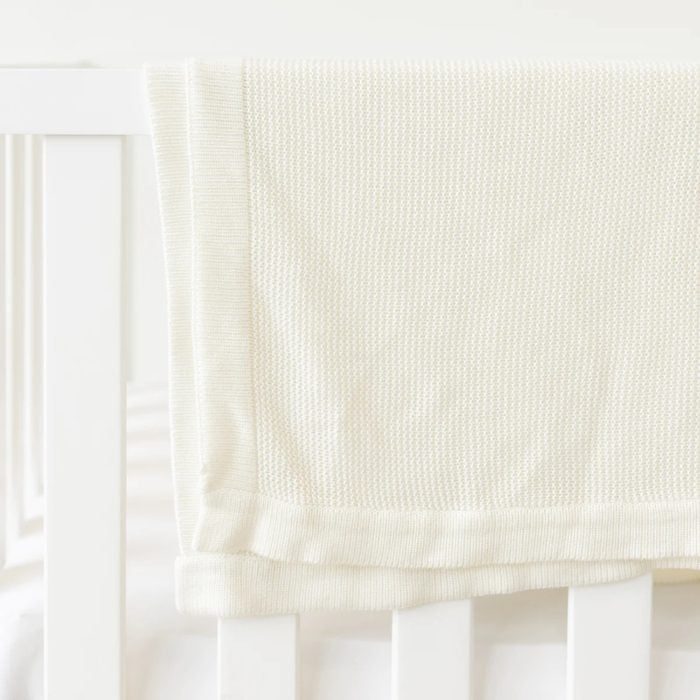 A cream-colored textured blanket rests draped over a white crib rail in a bright room.