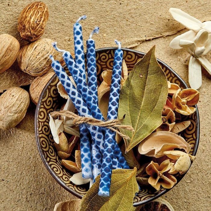 Blue patterned candles tied with twine rest in a decorative bowl filled with dried leaves, petals, and seeds on a rustic textured surface.